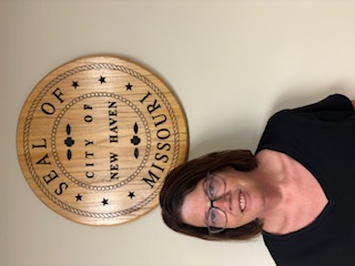 Woman seated indoors in front of a wall seal reading “SEAL OF THE CITY OF NEW HAVEN MISSOURI.”
